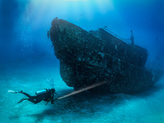 A scuba diver with a torch explores a sunken shipwreck at the seabed of the Maldives islands,...