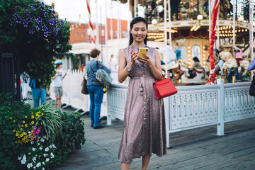 Attractive female tourist enjoying weekend leisure for visiting amusement park for recreation using cellphone for online messaging in social networks, millennial hipster girl installing mobile app