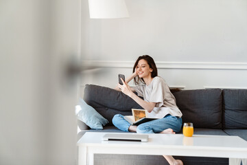 Photo of joyful beautiful girl using cellphone while sitting on couch