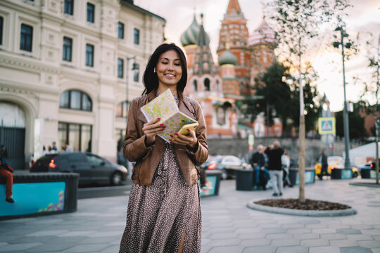 Happy Asian Tourist In Bluetooth Headphones Exploring Moscow City Using Cellphone Device And Paper Map For Tracking Gps Location During Travel Sightseeing, Cheerful Girl In Earbuds Enjoying Solo Trip