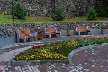 orange wooden bench with trash cans nearby, on stone path with flower beds with yellow and red flowers in park alleys under trees on green grass on cloudy day