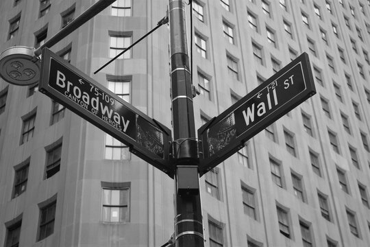 Low angle greyscale shot of a street sign with directions to Wall Street and Broadway in NYC