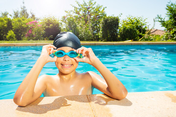 Happy little boy in swimming cap hold googles with hands on the border of outdoor pool look at...