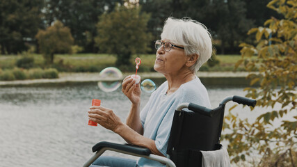 senior gray haired disabled woman in the wheelchair blowing soap bubbles near the river. High...