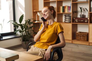 Image of happy redhead girl talking on cellphone while sitting at table