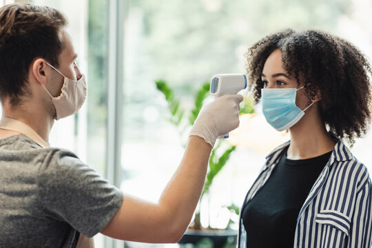 Young manager in protective mask and gloves checking temperature on client forehead at beauty salon