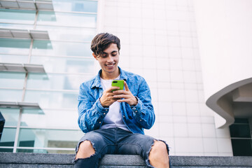 Cheerful caucasian male blogger checking income message on smartphone using 4G internet connection, smiling hipster guy millennial watching online video on mobile phone satisfied with leisure