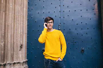 Smiling caucasian man in trendy outfit and eyewear standing on urban setting talking on mobile...