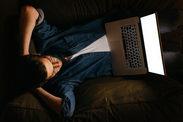 Young man in glasses laying on sofa at home, watching movies or online football game late at night,, relaxing after workday, has insomia, addicted to the Internet or TV series. Top view