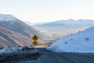 A winding road leads down the snow covered Crown Ranges, a slippery when wet road sign on the left warning drivers to take care during their travel. Arrow Junction, New Zealand.