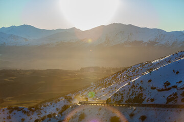 The sun starting to set over the snow covered mountains in the distance, viewed from the Crown ranges looking toward the road that leads down to Arrow Junction, lense flare from the sun.