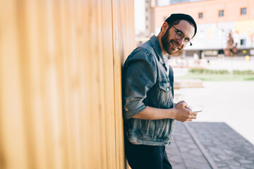 Half length portrait of happy Caucasian blogger in classic glasses for vision protection holding cellular technology and smiling at camera during sunny day in urban city, millennial hipster guy