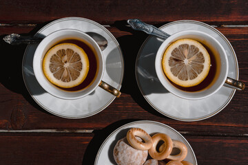 Beautiful white ceramic tableware for tea drinking. Two white cups of black tea with lemon on a wooden table, and delicious bagels and gingerbread in a saucer.