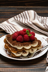 portion of Classic tiramisu dessert with raspberries on ceramic plate isolated on wooden background