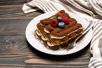portion of Classic tiramisu dessert with raspberries and blueberries on ceramic plate isolated on wooden background