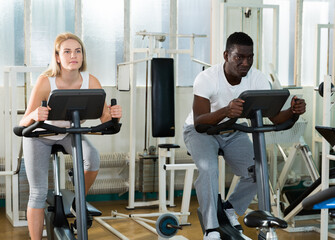 Fototapeta premium African man and Caucasian woman exercising on stationary bikes at gym