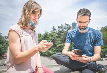 close up of  two young people with masks off  sitting in a park with serious looks to their phones. Social media and socialising during pandemics.