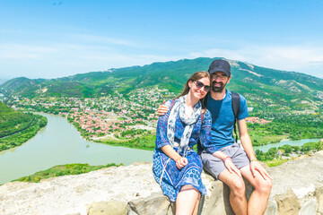 Couple sits together with scenic panorama mountais and Mtskheta view in the background and pose for a travel photo. Travel in summer and tourism in caucasus.