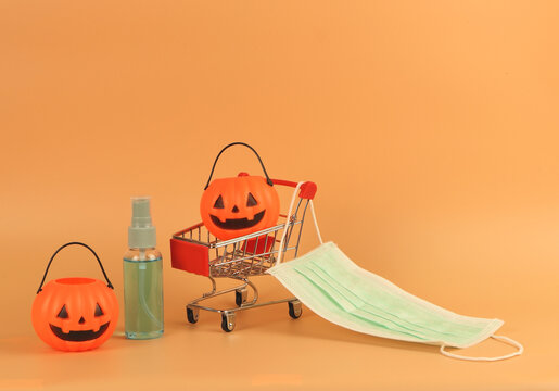 Halloween pumpkin on shopping cart with medical face mask and alcohol hand spray on orange background with copy space. Covid19 prevention , new normal and Halloween holiday concept.