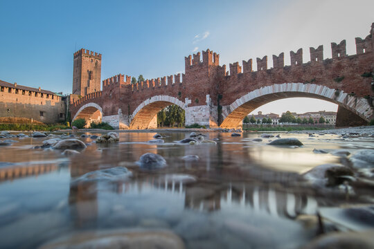 Low profile long exposure photo of a Castel vecchio bridge in Verona, Italy on a sunny day. Beautiful brick wall above the river of Adige on a blue sky