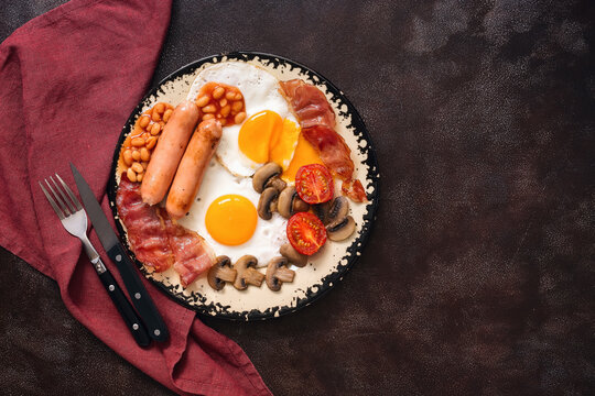 English breakfast: fried eggs with bacon, sausages, beans and mushrooms on a dark rustic background. Top view, flat lay, copy space.