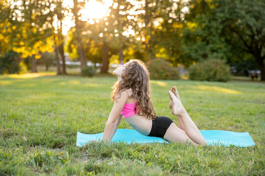 Full length profile image of a cute little girl with curly hair in sport wear doing yoga exercise on the roll mat in the park. Healthy lifestyle.