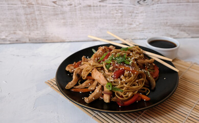 Udon noodle with vegetables and chicken fried on wok pan on the black plate with sticks and soy sauce on the sushi mat 