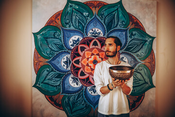 stylish bearded man holding a large Tibetan bowl on the background of a large color picture of a mandala