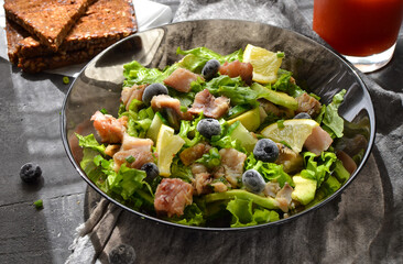 Salad with lettuce, fish, blueberries, lemon and avocado. Healthy lunch or brunch. Dark background. Linen gray napkin. Salad in a black glass plate. Backlight. Fish bowl with avocado.