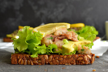 Dark bread sandwich with seeds. Sandwich with lettuce, avocado and fish. Healthy lunch or brunch. Dark background. Close-up.