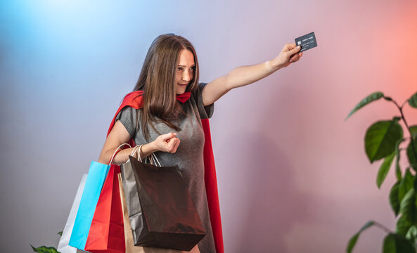 A Young Determined Woman In A Superhero Cape Is Holding Up Shopping Bags And Credit Card. Concept Of Sales And Shopping