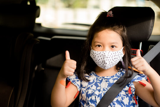 Smiling asian child girl thumbs up for good drive good trip and safe on the road.Sitting and buckle up in the car in the seat for children.Coronavirus covid-19.Little chinese girl wearing mask in car
