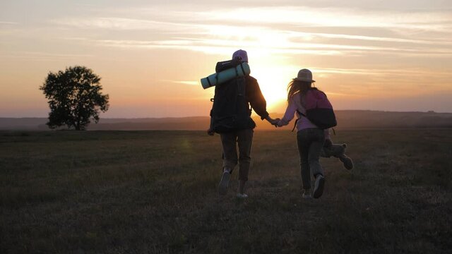 Mother and daughter with backpacks running through the meadow at sunset. Family tourism concept.