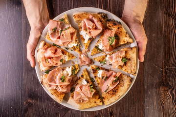 Mani che sorreggono una Pizza gourmet tagliata a spicchi con Mortadella, burrata pugliese, prezzemolo e granella di nocciole