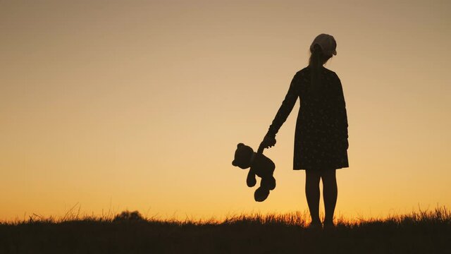 Little girl in dress holding teddy bear toy at sunset. Silhouette of a child with a toy. Concept of a child dream.