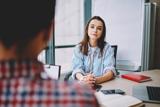 Selective focus on young woman listening attentively to male colleague during conversation in office, back view of man on frontage and smart casual female trainee have recruitment interview