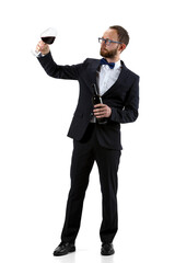 Fototapeta premium Trying red wine on. Portrait of male sommelier, wine steward or bar worker in white and black suit isolated over white background. Copyspace for ad. Concept of professional occupation, job.