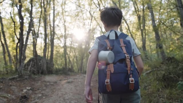 Happy boy tourist walking hiking through forest with tourist backpack with cup summer looks around at forest nature and animals slow motion. Lifestyle. Lens flare. Tourism. Childhood. Young discoverer