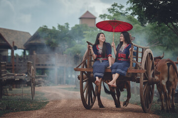 The way of life of Thai women from ancient times in traditional Thai costumes, sitting on a bullock cart or riding a cart. The backdrop is a beautiful ancient wooden house in Thailand.
