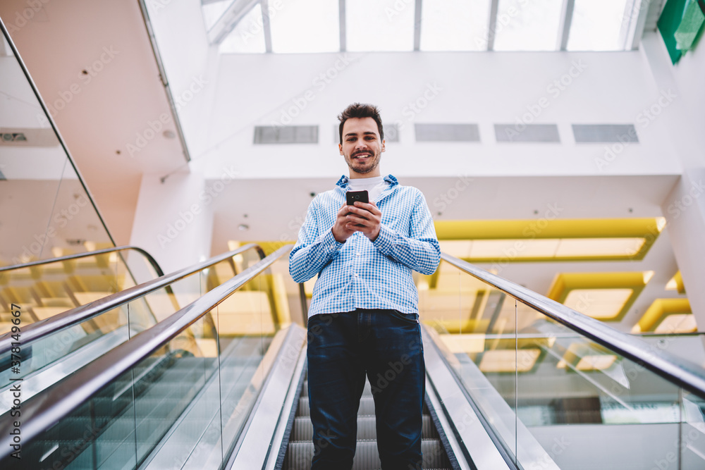 Poster half length portrait of cheerful caucasian man smiling at camera while going down on modern escalato