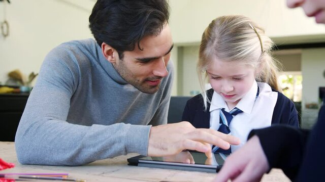Father helping son and daughter wearing school uniform with homework on digital tablet as they sit around kitchen table at home together - shot in slow motion
