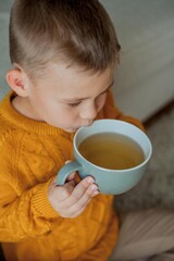A little cute boy in an orange sweater is drinking tea. Cozy portrait of a boy sitting at home. Fall.