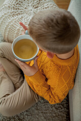 A little cute boy in an orange sweater is drinking tea. Cozy portrait of a boy sitting at home. Fall.