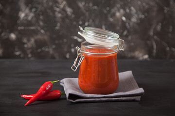 Harissa spicy condiment in a glass jar with a lid and chili on a black background.