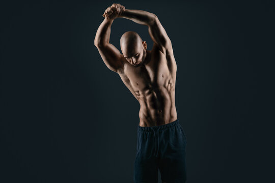 young bodybuilder look down isolated on black stretch his arms above his head while posing and showing trained muscles