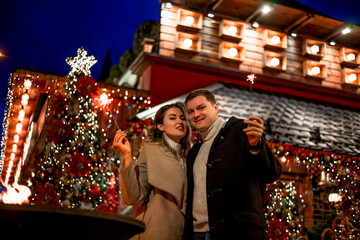 Couple in love relationship believe in meracle, kissing against the chrismas decoration outdoor in the eve, look at each other and holding sparklers
