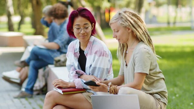 Young Asian female student with trendy pink hair discussing social media post on smartphone with Caucasian college groupmate while sitting together outdoors in park on summer day