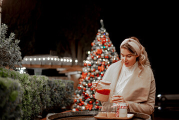 Happy woman holding warm drinklook at the new year tree posing against the festive decor and lights