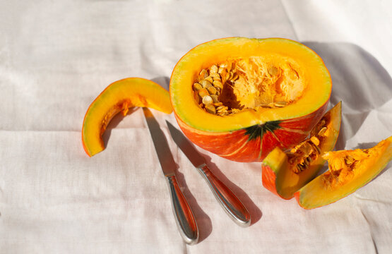  A ripe cut pumpkin and two steel kitchen knives on a woven napkin. Half pumpkin and wedges with seeds. Place for text on a light background.