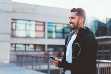 Happy Caucasian athlete 30 years old holding cellular in hands smiling at city urbanity, carefree male bodybuilder in electronic headphones for listening music enjoying time for outdoors workout